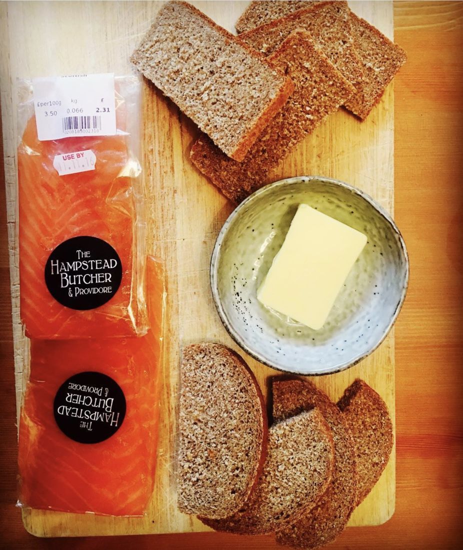Two packs of smoked salmon beside slices of dark wholemeal bread and a slab of butter in a stoneware bowl, all on a wooden board.