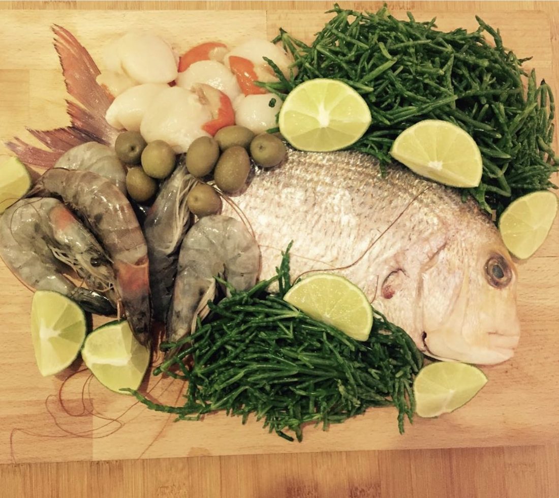 A whole sea bream on a wooden board, surrounded by raw prawns, scallops, green olives, lime wedges and a large pile of fresh samphire.