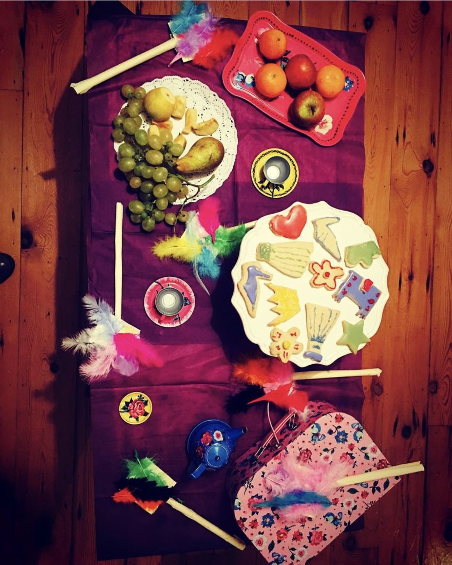 A party table on a purple cloth: a plate of biscuits iced by children, a bowl of grapes and pears, a tray of apples and satsumas, tea lights, a toy teapot and hand made feather wands.