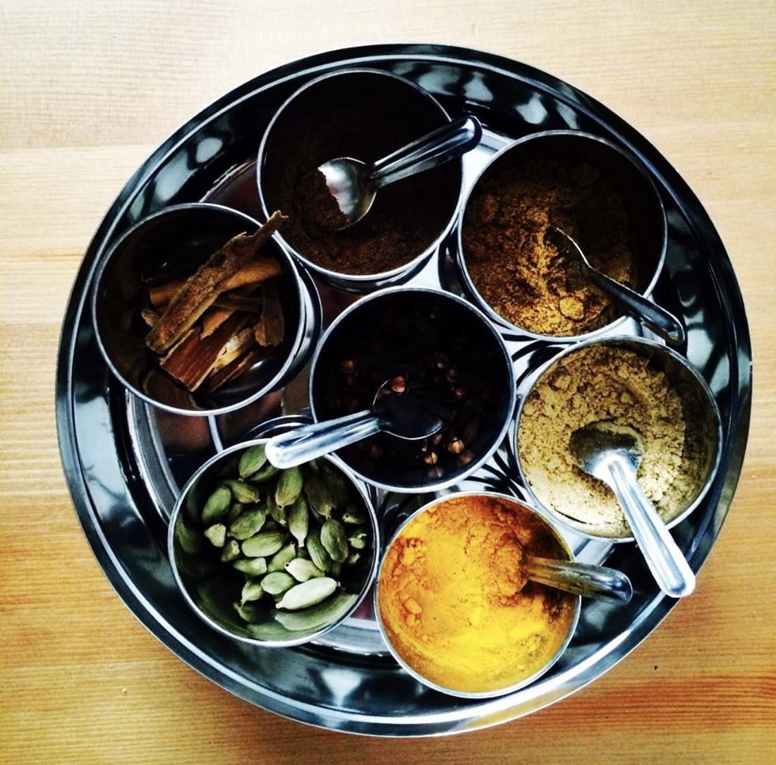An Indian spice tin from above, seven compartments holding turmeric, cardamom pods, cinnamon bark, black peppercorns, ground cumin and two ground spice blends, each with its own spoon.