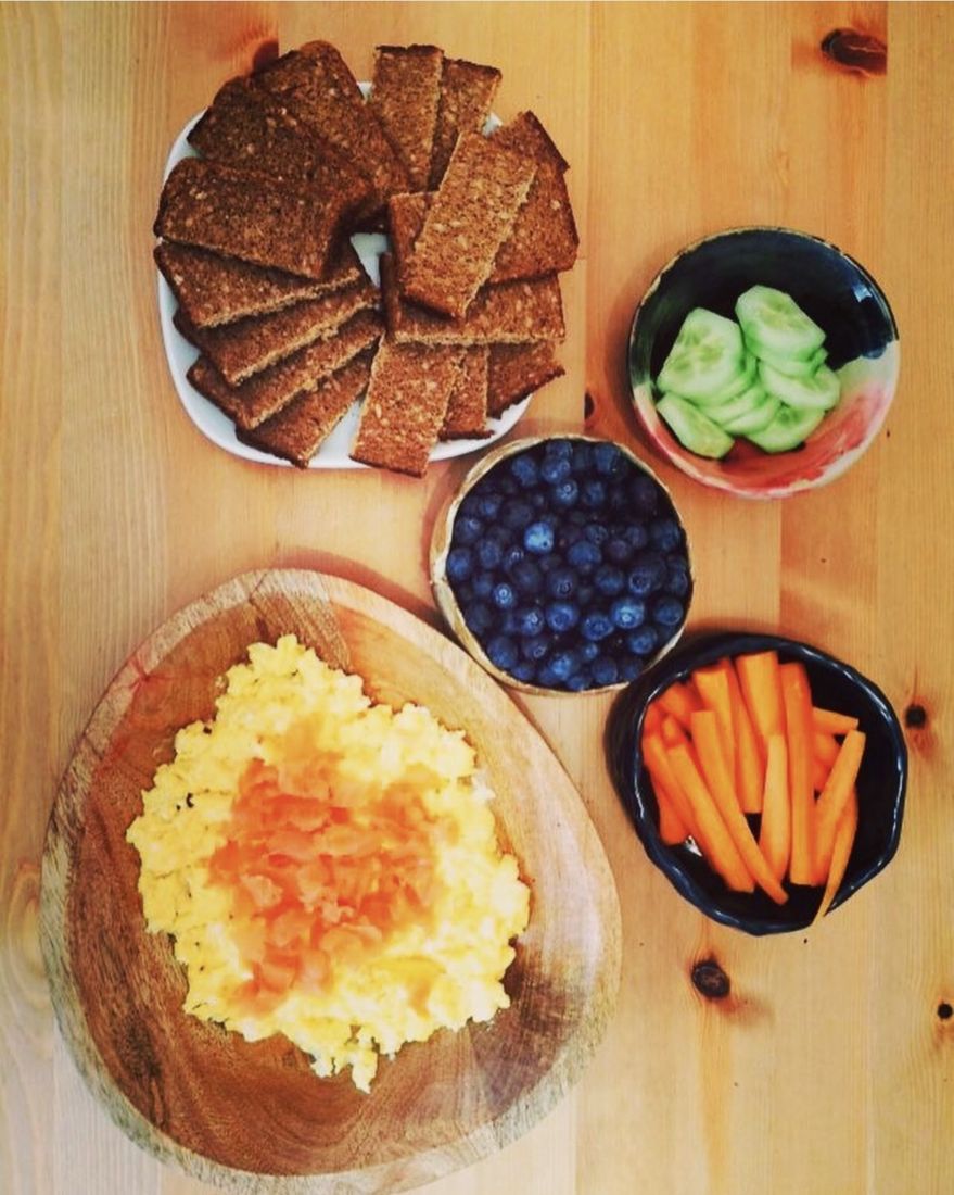 Scrambled egg on a round wooden board with a fan of wholemeal toast fingers, a bowl of blueberries, a bowl of cucumber slices and a bowl of carrot batons.