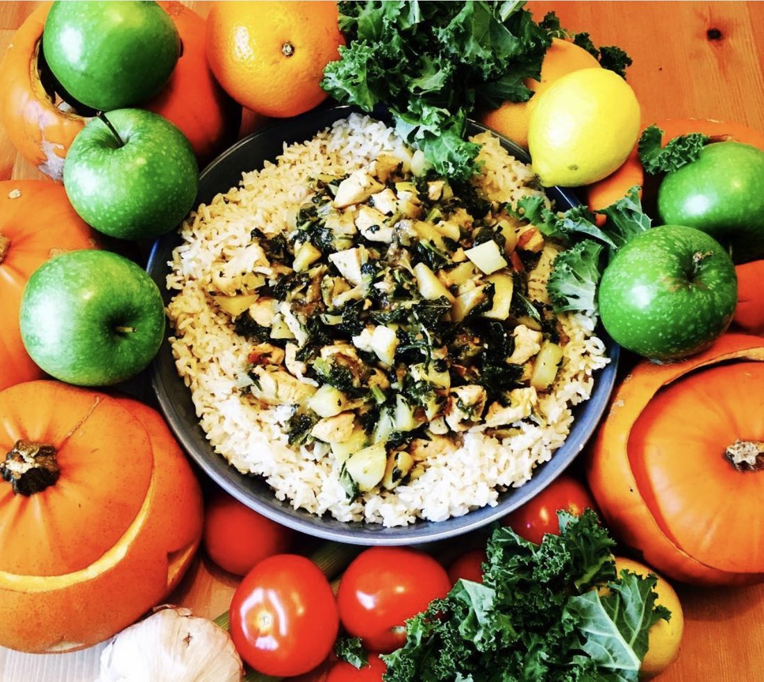 A bowl of rice topped with autumn chicken and vegetable stew, ringed by pumpkins, green apples, oranges, lemons, tomatoes, garlic and curly kale.
