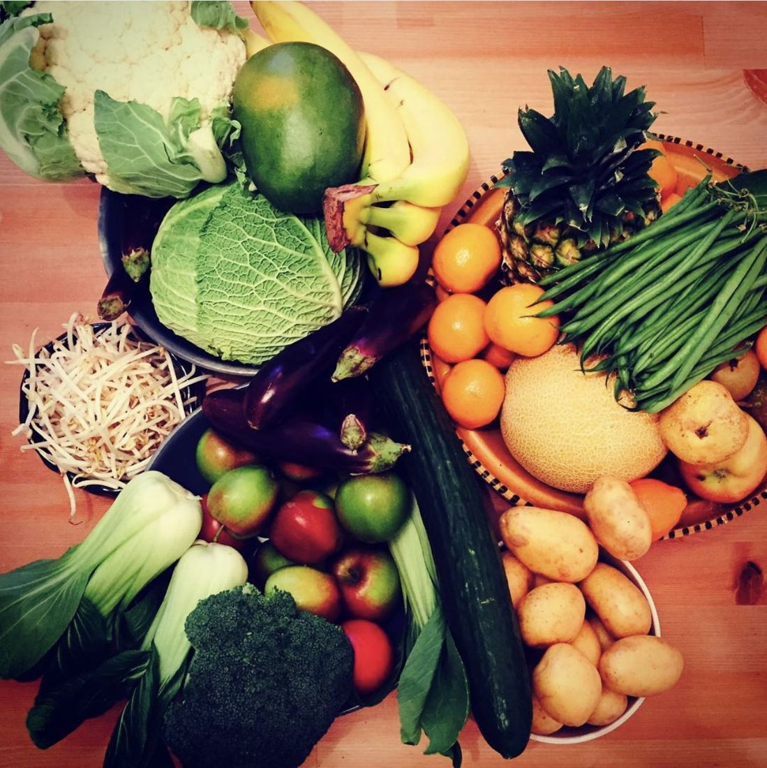 A week of fruit and vegetables laid out on a table: cauliflower, savoy cabbage, pak choi, aubergines, courgettes, beansprouts, broccoli, apples, bananas, a pineapple, satsumas, a melon, green beans and new potatoes.