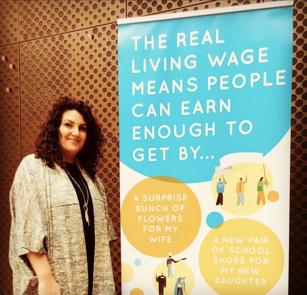 Yasmin standing beside a tall Living Wage Foundation banner at Hackney Town Hall. The banner reads: the real living wage means people can earn enough to get by, with illustrated circles saying a surprise bunch of flowers for my wife, and a new pair of school shoes for my new daughter.