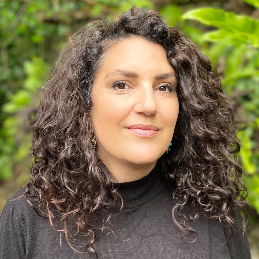 Yasmin A Darling, a mixed race woman with long dark curly hair, wearing a black roll neck, standing in front of green foliage and looking directly at the camera.